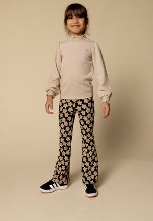 Jong meisje met een glimlach, gekleed in een beige lange mouw top, zwarte broek met beige bloemenpatroon, en zwart-witte sneakers, staande op een beige achtergrond.