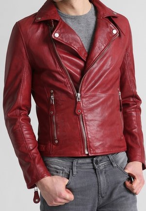 Mand iført en tætsiddende rød læder bikerjakke med sølv lynlåse over en grå rundhalset trøje og grå jeans, hænderne i lommerne.