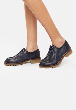 Chaussures en cuir bleu marine avec semelles en caoutchouc tan, présentant un design cousu et un système de laçage, mettant en valeur un bout rond et un profil bas.