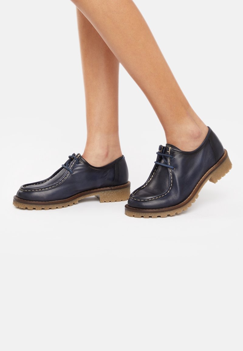 Chaussures en cuir bleu marine avec semelles en caoutchouc tan, présentant un design cousu et un système de laçage, mettant en valeur un bout rond et un profil bas.