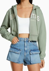 Mujer vestida con una chaqueta con cremallera corta de color verde claro, un top corto blanco y unos pantalones cortos de mezclilla azul de talle alto con grandes bolsillos frontales.