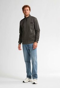 Giacca in pile verde scuro con zip, maniche lunghe e polsini a costine, indossata sopra jeans blu a gamba dritta e sneakers bianche.