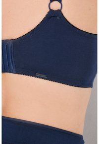 Sujetador azul marino con tejido suave, una silueta lisa, costuras decorativas y un acento metálico circular en la sujeción de la tira.