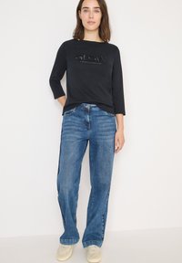 Musta puuvillainen pitkähihainen paita, jossa on "GLOW"-painatus, yhdistettynä vaaleansininhoisiin leveälahkeisiin farkkuihin, joiden pinta on sileä ja kangas denimtyyppistä.