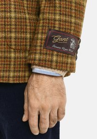 Nahaufnahme einer Hand eines Mannes, die eine braun karierte Wolljacke mit einem Etikett "Gant Genuine Virgin Wool" am Ärmel trägt.