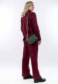 Abito su misura bordeaux con pantaloni a gamba larga, abbinato a una piccola borsa a tracolla verde. Il modello indossa stivali neri con suola spessa. Texture liscia.