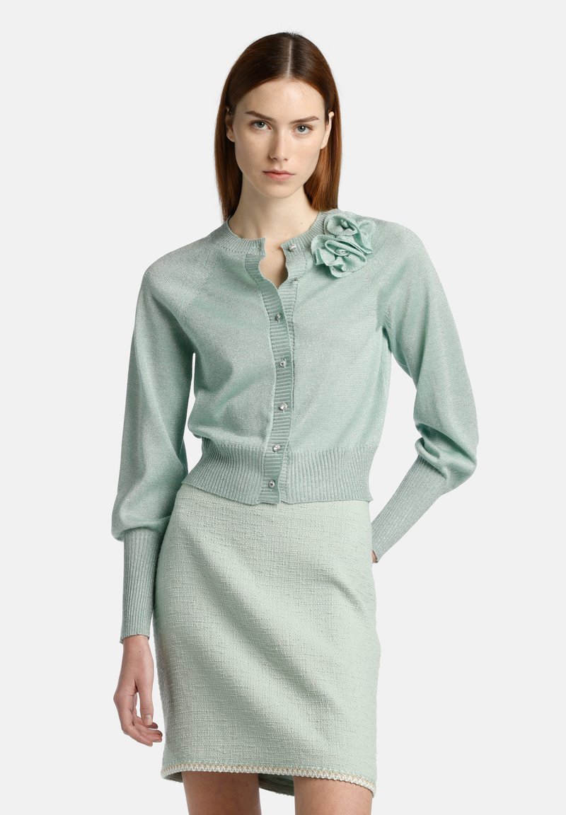 Mintgrüner Cardigan mit geripptem Saum und Bündchen, mit einem Blumenmotiv auf der Schulter und Knopfverschluss. Getragen mit einem passenden Rock.