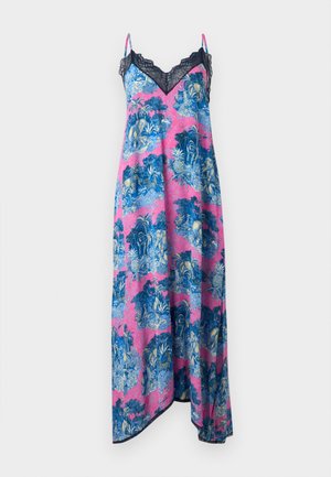 Kobieta w różowo-niebieskiej sukni maxi w kwiaty z granatową koronkową lamówką oraz czarnych butach do połowy łydki z łańcuchowym detalem, stojąca na białym tle.