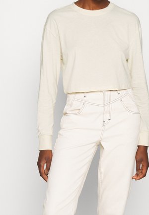 Osoba ubrana w kremowy, długi rękaw, krótki top oraz białe, jeansy z wysokim stanem, z widocznymi przednimi kieszeniami i detalami przeszyć.