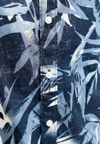 Közeli kép egy sötétkék ingről, amelyen absztrakt, világoskék és fehér ecsetvonásminták láthatók, valamint három fehér gomb.