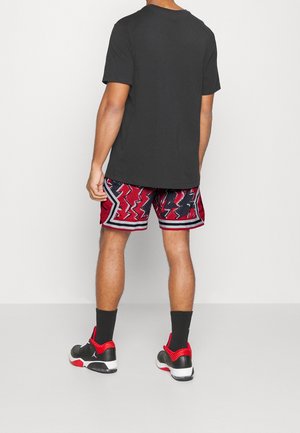 Schwarzes kurzärmeliges T-Shirt kombiniert mit roten gemusterten Shorts mit Zickzack-Designs, grauen Akzenten und schwarzen Sneakers mit roten Details.
