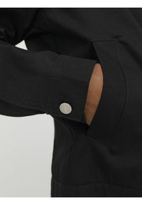 Poignet de veste en tissu noir avec un bouton-pression en métal argenté, montrant une texture lisse et une couture plate, avec une poche latérale visible.