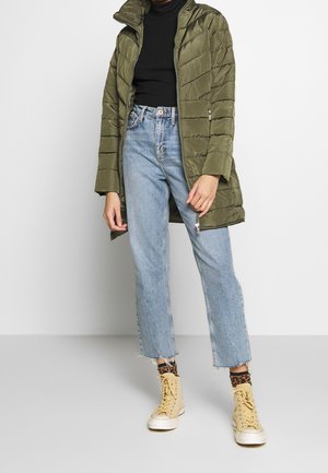 Person iført olivengrøn quiltet jakke, sort top, lyseblå jeans, leopardmønstrede sokker og beige høje sneakers på gråt gulv.