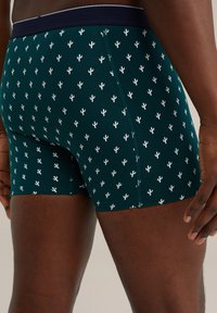 Sötétzöld anyagú boxeralsó, amelyen ismétlődő fehér kaktuszminta található, elasztikus derékkal és laposvarrásos varrással a kényelem érdekében.