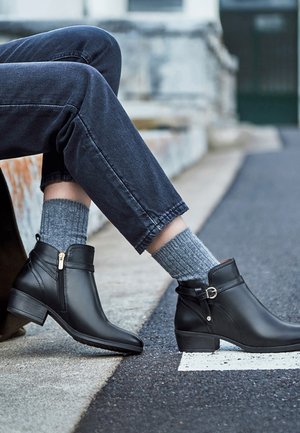 Osoba nosiaca čierne kožené členkové topánky so zipsami a prackami, sivé ponožky a tmavé džínsy, sediaca na obrubníku pri ulici.