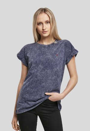 Femme blonde portant un t-shirt bleu délavé à manches retroussées avec un petit motif main et cœur sur la poitrine, posant avec une main dans la poche.