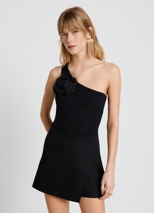 Femme portant une robe noire à une épaule avec une grande fleur en tissu sur la bretelle et des boucles d'oreilles pendantes en forme de feuille dorée.