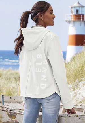 Woman with brown hair in ponytail wearing white hooded jacket and light blue jeans near beach with lighthouse in background.