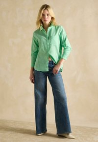 Groene knoopshirt gemaakt van lichtgewicht stof, met een zak en opgerolde mouwen, gecombineerd met uitlopende blauwe denim jeans.