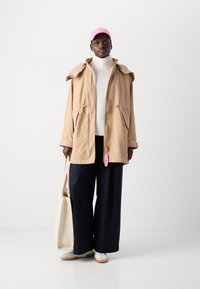 Lys beige parkajakke med avtakbar hette, kremfarget turtleneck, marineblå vide bukser, rosa caps og hvite joggesko. Beige tote bag.
