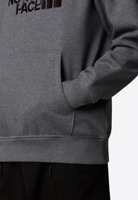 Sudadera gris con puños y dobladillo acanalados, que presenta un bolsillo delantero y un logo negro en la zona del pecho. Tejido suave y texturizado.