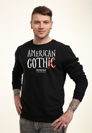 Man met tatoeages die een zwart sweatshirt draagt met de tekst "American GothX Wednesday", staand met zijn handen op zijn heupen tegen een effen lichte achtergrond.