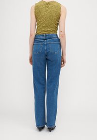 Top sin mangas de color verde oliva con textura, combinado con jeans rectos de tiro alto en color azul; bolsillos traseros y costuras visibles en los jeans.