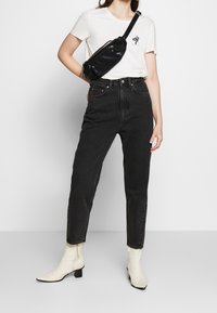 Jeans in denim nero con vita alta e vestibilità rilassata, abbinati a una maglietta bianca con una piccola grafica e una borsa a tracolla nera. Stivaletti bianchi alla caviglia.