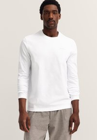 Mann in einem weißen longsleeve Hemd und hellgrauen Hosen, stehend vor einem einfarbigen beigen Hintergrund, mit Blick nach vorne.