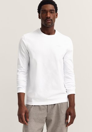 Mann in einem weißen longsleeve Hemd und hellgrauen Hosen, stehend vor einem einfarbigen beigen Hintergrund, mit Blick nach vorne.