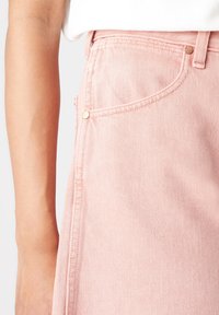 Primer plano de una persona que lleva pantalones de mezclilla rosa claro con un bolsillo delantero visible, un ojal para el cinturón y parte de una camisa blanca.