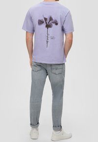 Camiseta de algodón lavanda con un diseño floral negro en la parte posterior. Combinada con jeans grises y zapatillas blancas. Presenta un cuello redondo clásico.