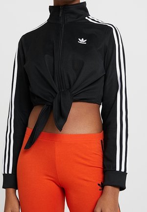 Czarna krótka kurtka sportowa Adidas z białymi paskami i wiązaniem z przodu, zestawiona z jaskrawo pomarańczowymi legginsami Adidas.