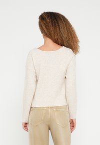 Vrouw met krullend haar, gekleed in een geribd, crèmekleurig top met lange mouwen en beige broek, van achteren getoond tegen een effen achtergrond.