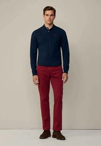 Mann in navyblauem Langarm-Polo, roten Hosen und braunen Schnürschuhen, der vor einem schlichten beigen Hintergrund steht.