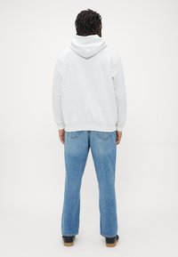 Person med mørkt flettet hår som har på seg en lys grå hettegenser, blå jeans og svarte sko, som står vendt bort fra kameraet mot en ensfarget bakgrunn.