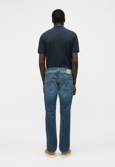 Άνδρας που φοράει σκούρο μπλε πόλο, μπλε τζιν με πίσω τσέπες και καφέ σουέτ παπούτσια, στέκεται γυρισμένος με την πλάτη προς μια απλή φωτεινή επιφάνεια.