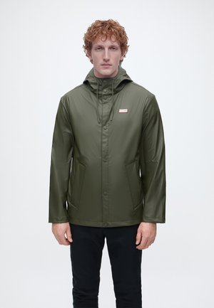 Bărbat cu păr creț roșcat, purtând o geacă de ploaie verde măsliniu cu glugă și pantaloni negri, stând în fața unui fundal alb simplu.