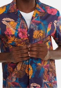 Camicia a maniche corte con bottoni, caratterizzata da una stampa tropicale multicolore su uno sfondo viola scuro, con foglie di palma e uccelli ben visibili.