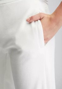 Weiße Hose mit glatter Textur, ausgestattet mit einer Seitentasche und einer lockeren Passform, abgebildet mit einer Hand, die in die Tasche gesteckt ist.