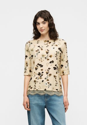 Woman with wavy dark hair wearing a beige floral top with lace trim and blue jeans, standing against a plain light background.