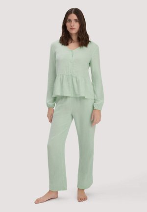 Junge Frau steht barfuß und trägt eine hellgrüne, strukturierte Bluse mit langem Ärmel und Peplum sowie passende weite Hosen.