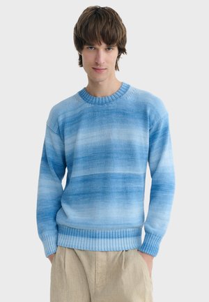 Jeune homme portant un pull tricoté à dégradé bleu clair et un pantalon beige, les mains dans les poches, debout devant un fond uni.
