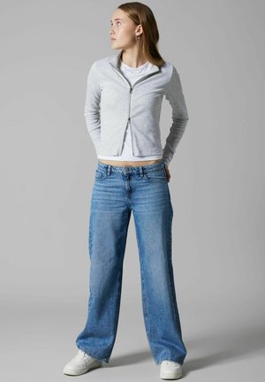 Grå zip-up jakke over en hvid t-shirt, parret med brede blå jeans og hvide sneakers. Glat stof, afslappet design.