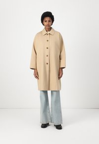 Cappotto beige oversize con colletto, sette bottoni grandi e due tasche laterali. Abbinato a jeans a campana azzurro chiaro e scarpe nere.