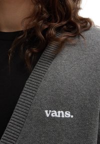 Grå cardigan med ribbad kant och V-ringning. Har en vit broderad "vans."-logotyp på vänster bröstområde. Slät textur.