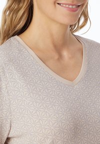 V-Ausschnitt-Oberteil in hellbeige mit einem strukturierten Muster überlappender Blattformen in Blau. Weicher Stoff mit subtilem Glanz.