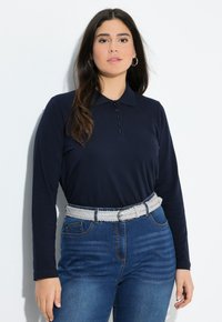 Marineblaues Langarm-Polohemd mit Kragen und sechs Knöpfen, kombiniert mit hochgeschnittenen blauen Jeans und einem geflochtenen weißen Gürtel.