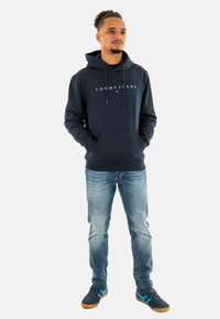 Marine hoodie met een voorzak, groene "TOMMY JEANS" logo, gecombineerd met lichtblauwe jeans en donkere sneakers met blauwe accenten.