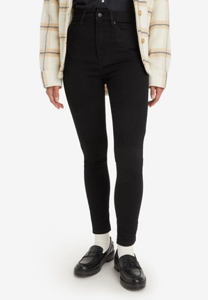 Black high-waisted skinny jeans with a smooth texture, paired with black loafers and white socks, under a cream and plaid jacket.